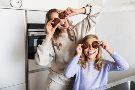 Image of a happy young woman with her little sister indoors at home kitchen covering eyes with cookies.の写真素材
