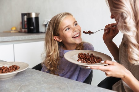 Image of two happy girls sisters at kitchen indoors eat have a breakfast together.
