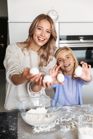 Image of a happy young woman with her little sister indoors at home kitchen cooking with flour and eggs.の写真素材