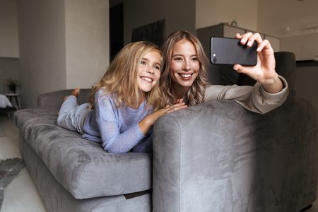 Image of a happy young woman with her little sister indoors at home take a selfie by mobile phone.の写真素材