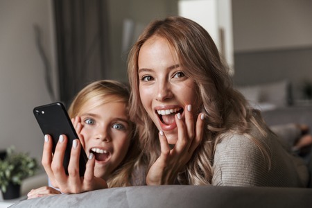 Image of a happy young woman with her little sister indoors at home using mobile phone.