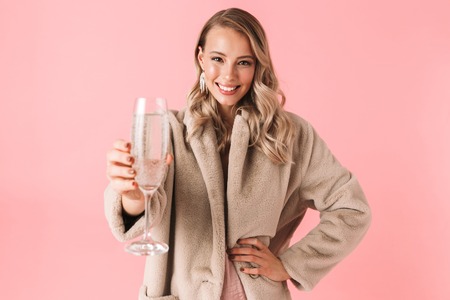 Photo of a beautiful emotional young pretty woman posing isolated over pink wall background holding glass with alcohol drink.の写真素材