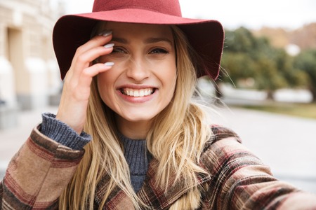 Beautiful young stylish blonde woman wearing a coat walking outdoors, taking a selfie with mobile phoneの写真素材