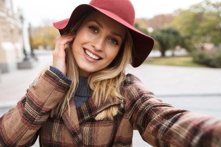 Beautiful young stylish blonde woman wearing a coat walking outdoors, taking a selfie with mobile phoneの写真素材