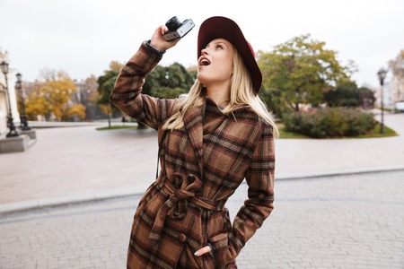 Beautiful young stylish blonde woman wearing a coat walking outdoors, taking pictures with photo cameraの写真素材