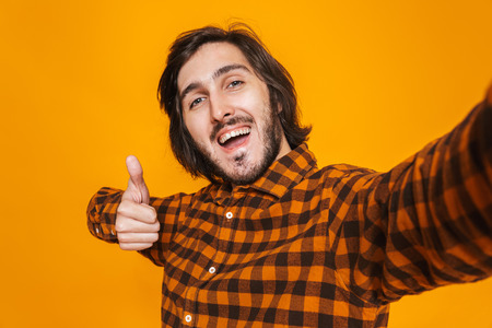 Portrait of smiling man wearing plaid shirt rejoicing and taking selfie photo while standing isolated over yellow background in studioの写真素材