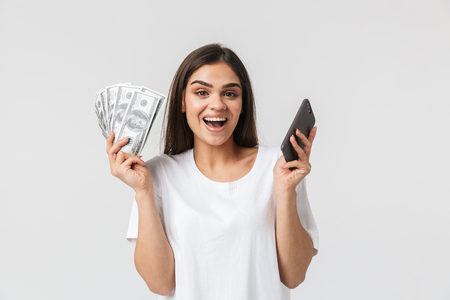 Close up portrait of a happy pretty young woman casualy dressed isolated over white, showing money banknotes, using mobile phoneの写真素材