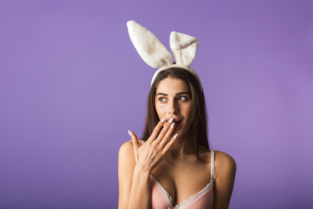 Beautiful sensual brunette woman wearing lingerie and bunny ears standing isolated over violet backgroundの写真素材