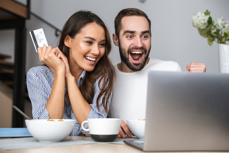Happy multiethnic couple having breakfast at the kitchen, looking at laptop computer, showing credit cardの写真素材