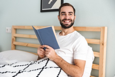 Handsome young man laying in bed, working on laptop computer, reading a bookの写真素材