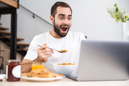 Handsome young man having breakfast while sitting at the kitchen, usinh laptop computerの写真素材