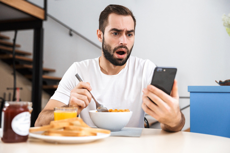 Handsome shocked young man having breakfast while sitting at the kitchen, holding mobile phone