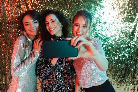 Three cheerful beautiful women wearing bright clothes having a party over sparkling background, taking a selfieの写真素材