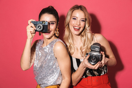 Image of two adorable girls 20s in stylish outfit holding and photographing on retro camera isolated...