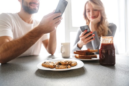 Image of amazing young loving couple sitting at the kitchen have a breakfast using mobile phones.