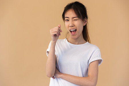 Photo of cute korean woman wearing basic t-shirt smiling and winking at you isolated over beige background in studioの写真素材