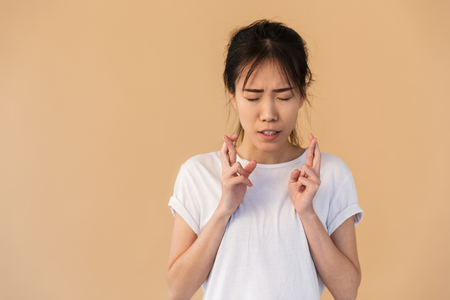 Portrait of concentrated asian woman wearing basic t-shirt begging god please with fingers crossed isolated over beige background in studioの写真素材