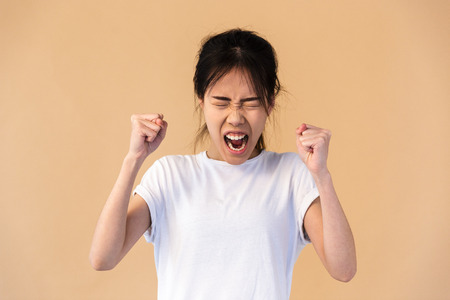 Photo of emotional korean woman wearing basic t-shirt smiling and clenching fists isolated over beige background in studioの写真素材