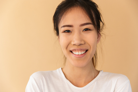 Portrait closeup of optimistic asian woman wearing basic t-shirt laughing and looking at camera isolated over beige background in studioの写真素材