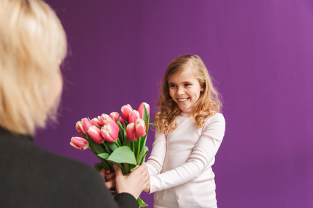 Portrait of a smiling little girl holding bouquet of tulips isolated over violet background, giving to a womanの写真素材