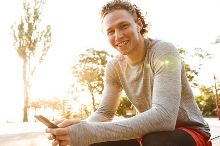 Confident sportsman wearing earphones sitting outdoors, using mobile phone
