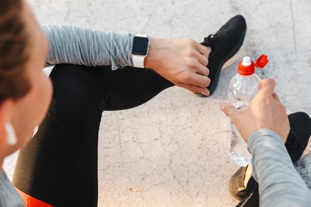 Top view of a young sportsman wearing earphones sitting outdoors, holding bottle of waterの写真素材