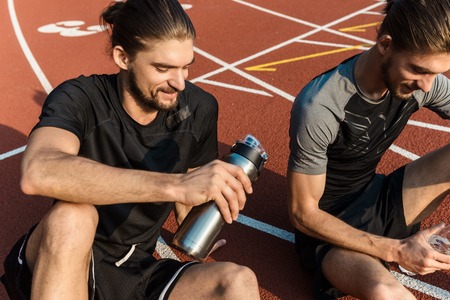 Two twin brothers resting after workout while sitting on a stadium, drinking warerの写真素材