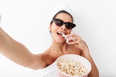 Cheerful young woman sitting in bed after shower wrapped in blanket, wearing sunglasses, eating popcornの写真素材