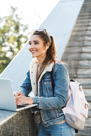 Smiling young woman wearing jacket standing at the stairs outdoors, using laptop computerの写真素材