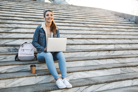 Smiling young woman wearing jacket sitting on stairs outdoors, using laptop computerの写真素材