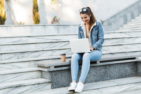 Smiling young woman wearing jacket sitting on a bench outdoors, using laptop computerの写真素材