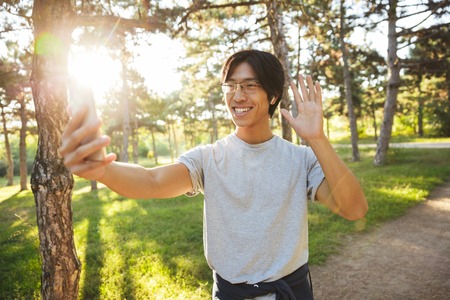 Smiling asian athlete man wearing sportswear taking a selfie while standing at the park, waving hand