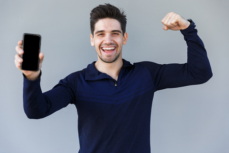 Cheerful young man showing blank screen mobile phone while standing isolated over grayの写真素材
