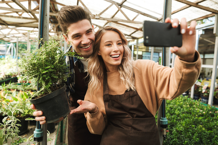 Photo of emotional happy young two colleagues gardeners at the workspace over plants take a selfie by mobile phone.の写真素材
