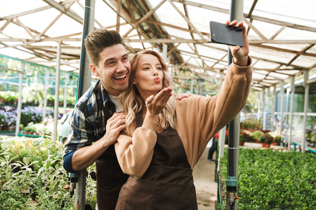 Image of a happy young loving couple gardeners at the workspace over plants take a selfie by mobile phone.の写真素材