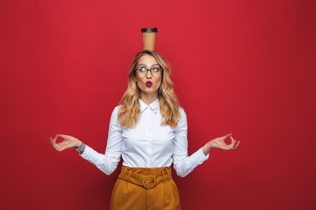 Beautiful funny young blonde woman standing isolated over red background, balancing, holding coffee cupの写真素材