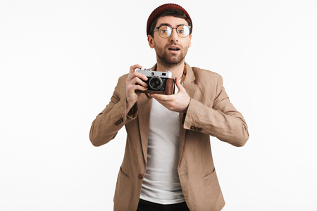 Photo of european man 30s wearing hipster hat rejoicing while holding and photographing on retro camera isolated over white backgroundの写真素材