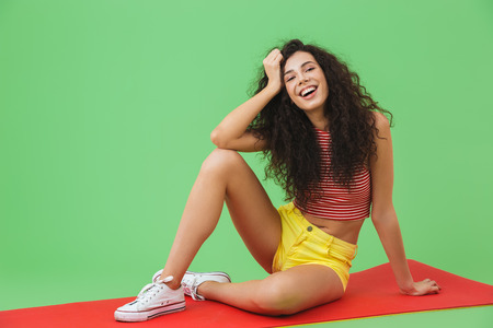 Photo of european fitness woman 20s smiling and sitting on yoga mat during aerobics against green...