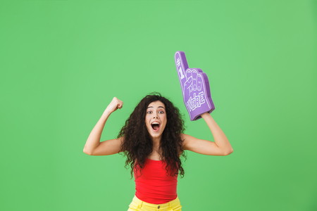 Portrait of modern woman 20s rejoicing and holding number one fan glove while standing against green wallの写真素材