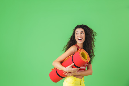 Photo of european woman 20s smiling and carrying yoga mat during workout against green wallの写真素材