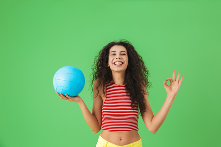 Image of joyful woman 20s wearing summer clothes smiling and holding volley ball while standing against green backgroundの写真素材