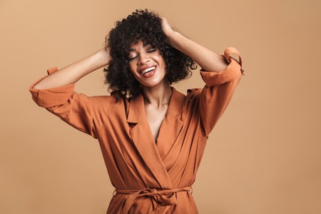 Smiling pretty african woman posing with closed eyes and holding her head over brown backgroundの写真素材