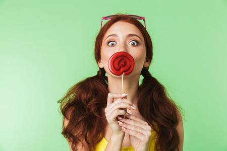 Image of a shocked beautiful excited young redhead girl posing isolated over green wall background with candy.の写真素材