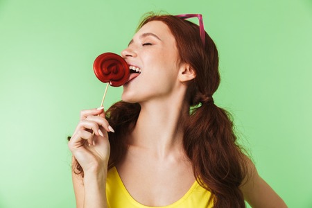 Image of a beautiful excited young redhead girl posing isolated over green wall background with candy.の写真素材