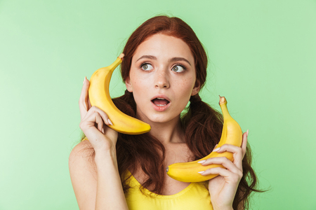 Image of a beautiful shocked excited young redhead girl posing isolated over green wall background with bananas fruits.の写真素材