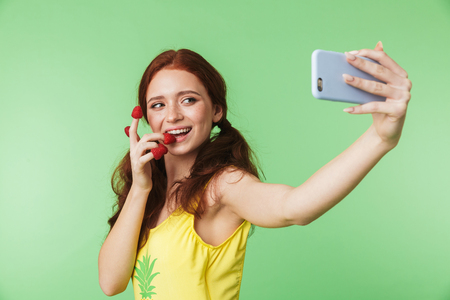 Image of a beautiful emotional young redhead girl posing isolated over green wall background with raspberry take selfie by phone.の写真素材