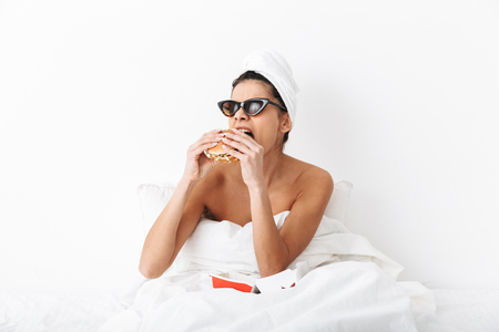 Image of a beautiful emotional woman with towel on head lies in bed under blanket isolated over white wall background wearing sunglasses eat burger.の写真素材