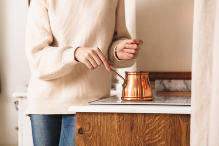 Image of caucasian brunette woman 30s brewing coffee in stainless steel turkish coffee pot on plate in kitchenの写真素材