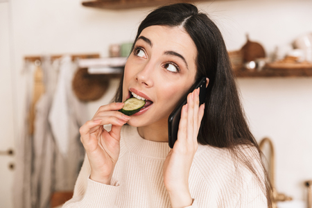 Picture of adorable brunette woman 30s eating cucumber while holding and talking on smartphone in stylish kitchen at homeの写真素材