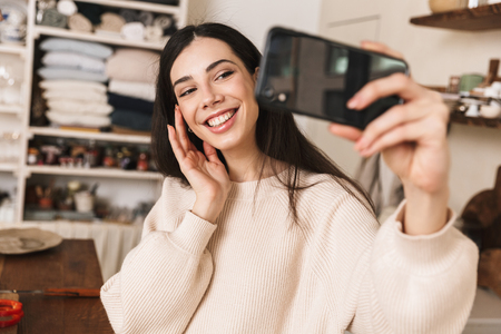 Portrait of satisfied brunette woman 30s taking selfie photo on smartphone while cooking green salad with vegetables in stylish kitchen at homeの写真素材